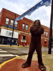 Bigfoot stands on a street corner in front of a banner reading "Ellwood City." His arms are crossed, he's looking up, and he's ready to do some sight-seeing.