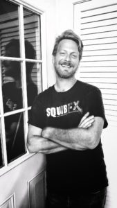 Black and white image of Steve Tolin wearing a Squib FX T-shirt leaning against the glass pane of the door of the Tolin FX studio in Beechview