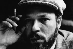 Black and White image of playwright August Wilson wearing his trademark cap, looking pensive and holding a pen.